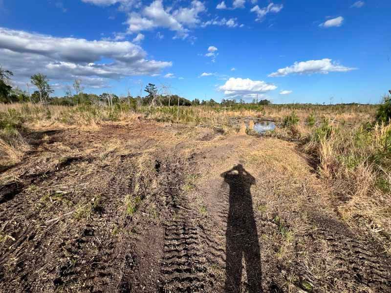 Rough dirt track through dry grassland under a bright blue sky, with a long human shadow in the foreground