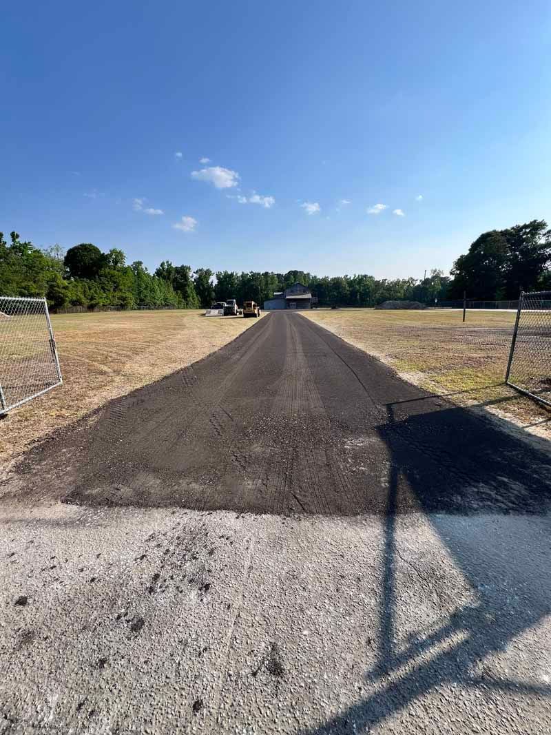 Gravel driveway leading to a small building under a clear blue sky, flanked by dry fields and trees