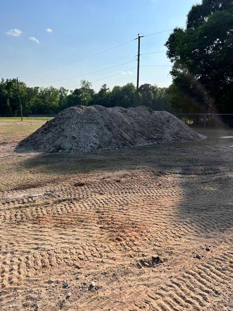 A large gravel pile on a dirt lot with tire tracks, trees, and a utility pole in the background.