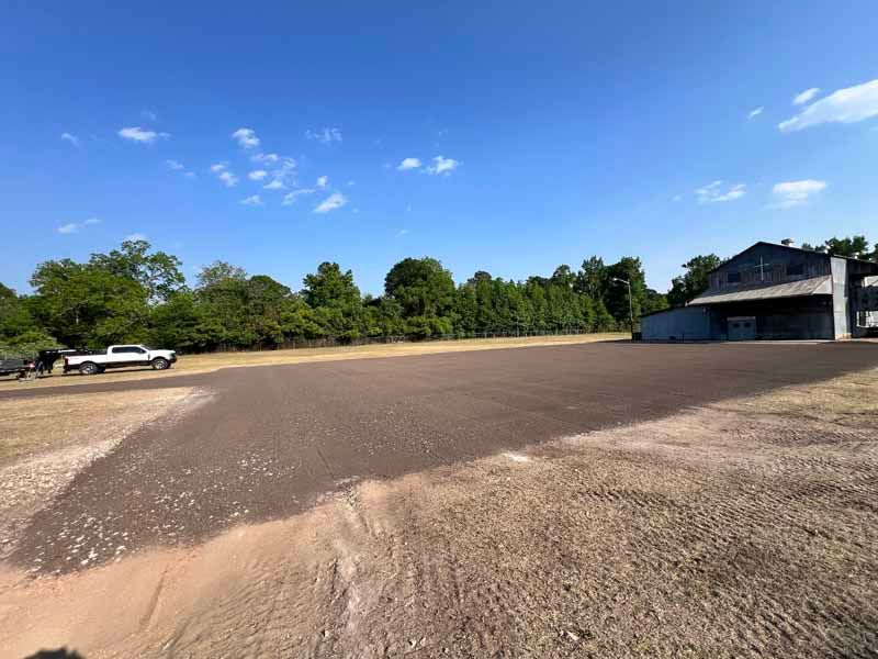 Wide gravel lot beside a small building, with a white truck and trees under a blue sky