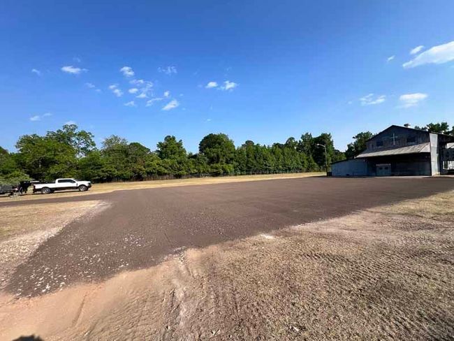 Wide gravel lot beside a small building, with a white truck and trees under a blue sky