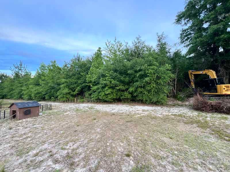 Gravel lot beside dense green trees, with a small shed on the left and an excavator on the right.