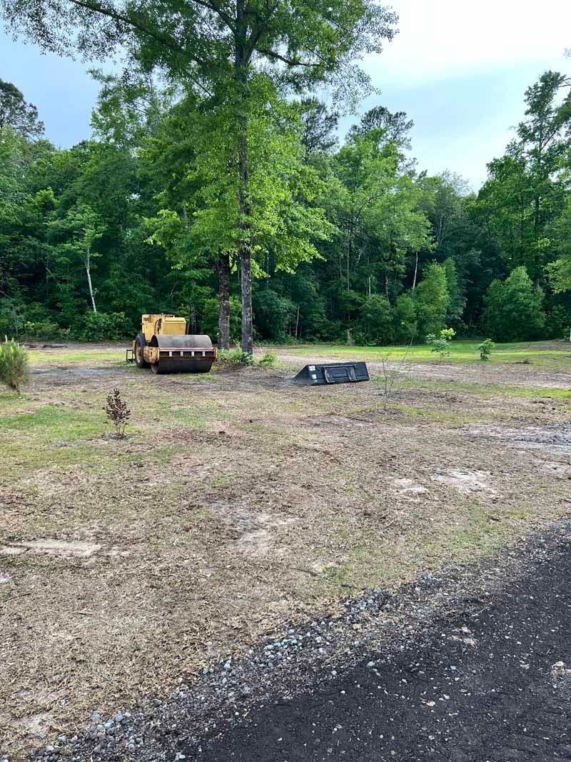 Cleared dirt lot beside a wooded area with a yellow excavator and a small black dumpster.