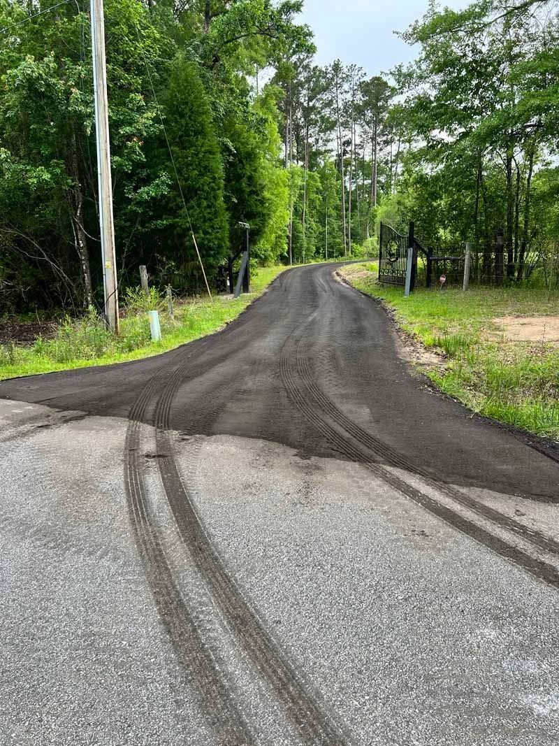 Narrow gravel road with fresh dark asphalt patch winding through a green wooded area.
