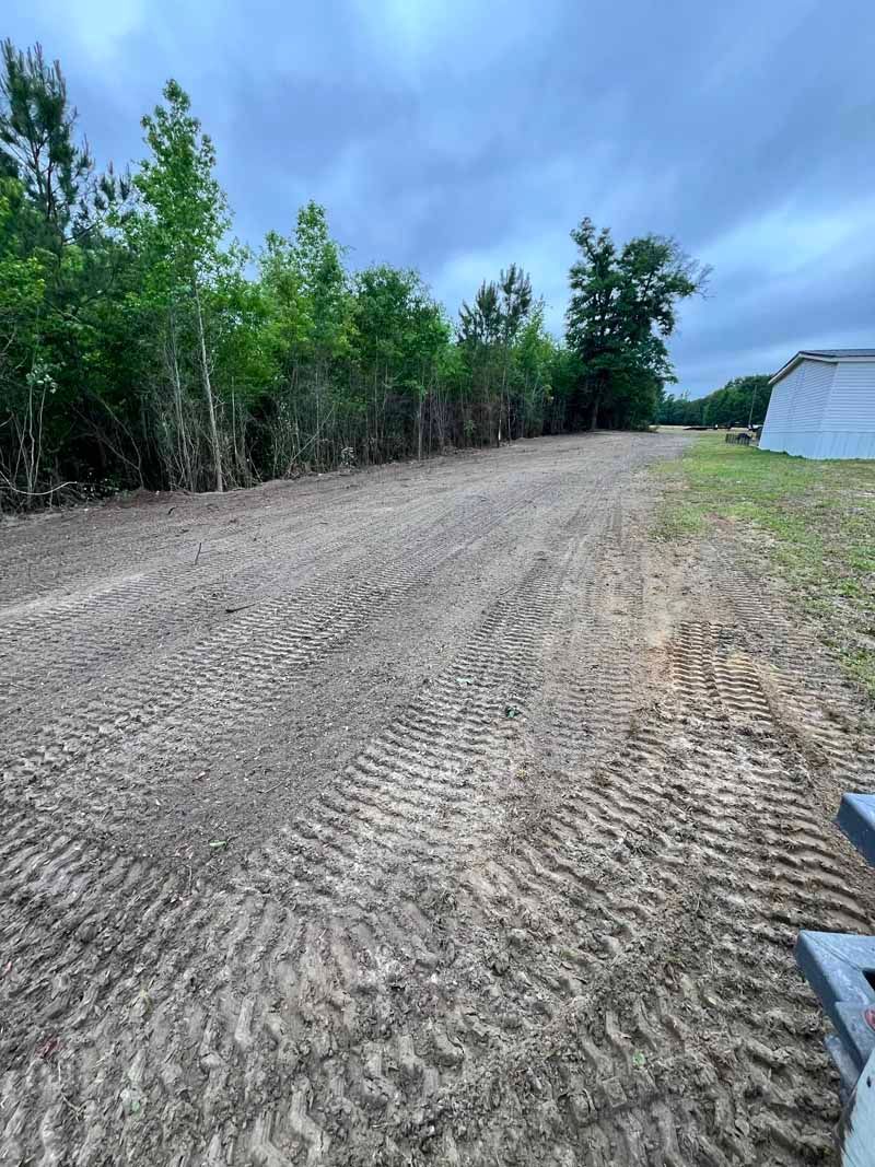 Graded dirt road beside green trees under a cloudy sky, with a white building on the right.