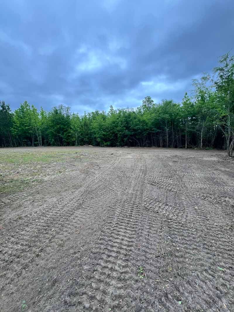 Cleared dirt lot bordered by green trees under a cloudy blue sky