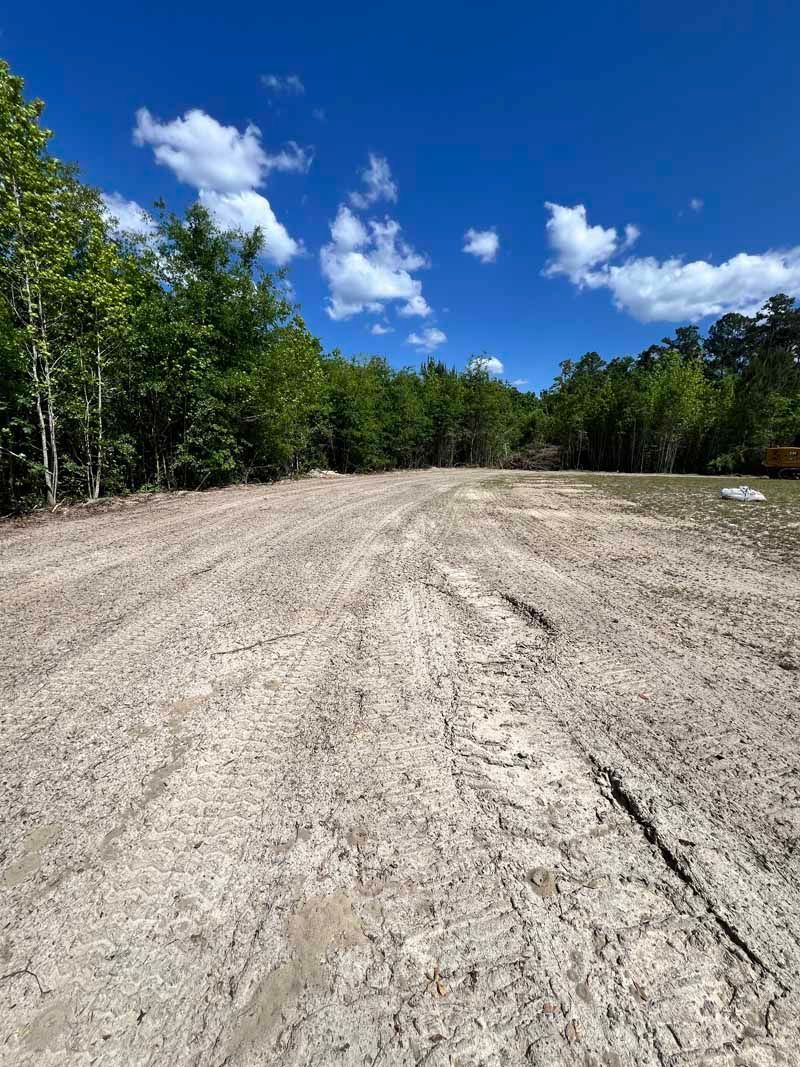 Wide dirt clearing beside green trees under a bright blue sky with scattered white clouds