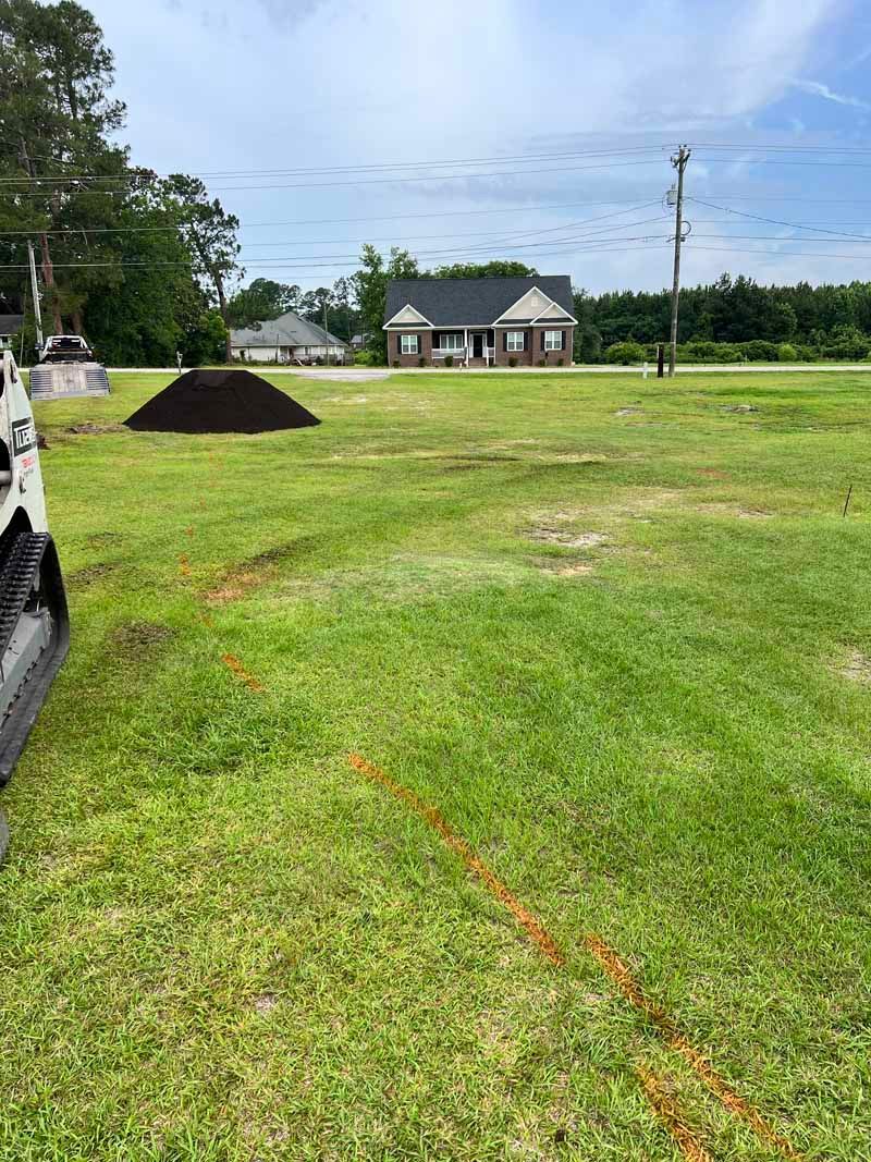 Grassy lot with a dirt pile, utility pole, and houses under a bright blue sky