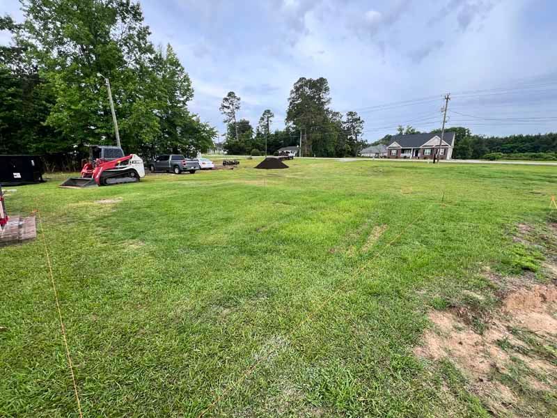 Wide grassy field with parked vehicles, trees, and a house under a cloudy sky