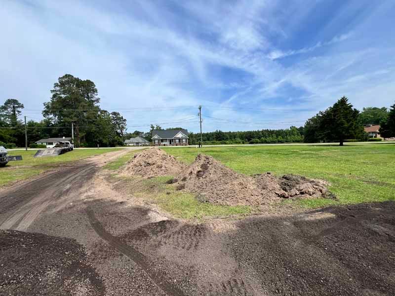 Rural dirt road beside a grassy field with a mound of soil under a blue sky