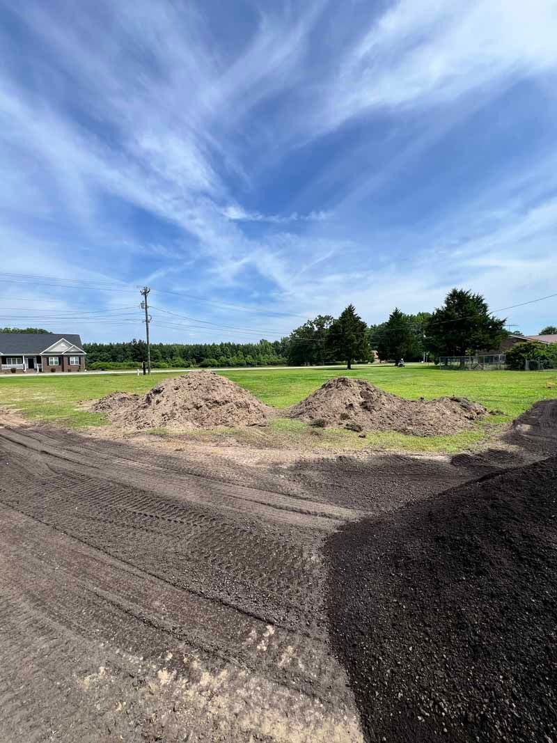 Open dirt lot with gravel piles, grassy field, trees, and a blue sky with wispy clouds