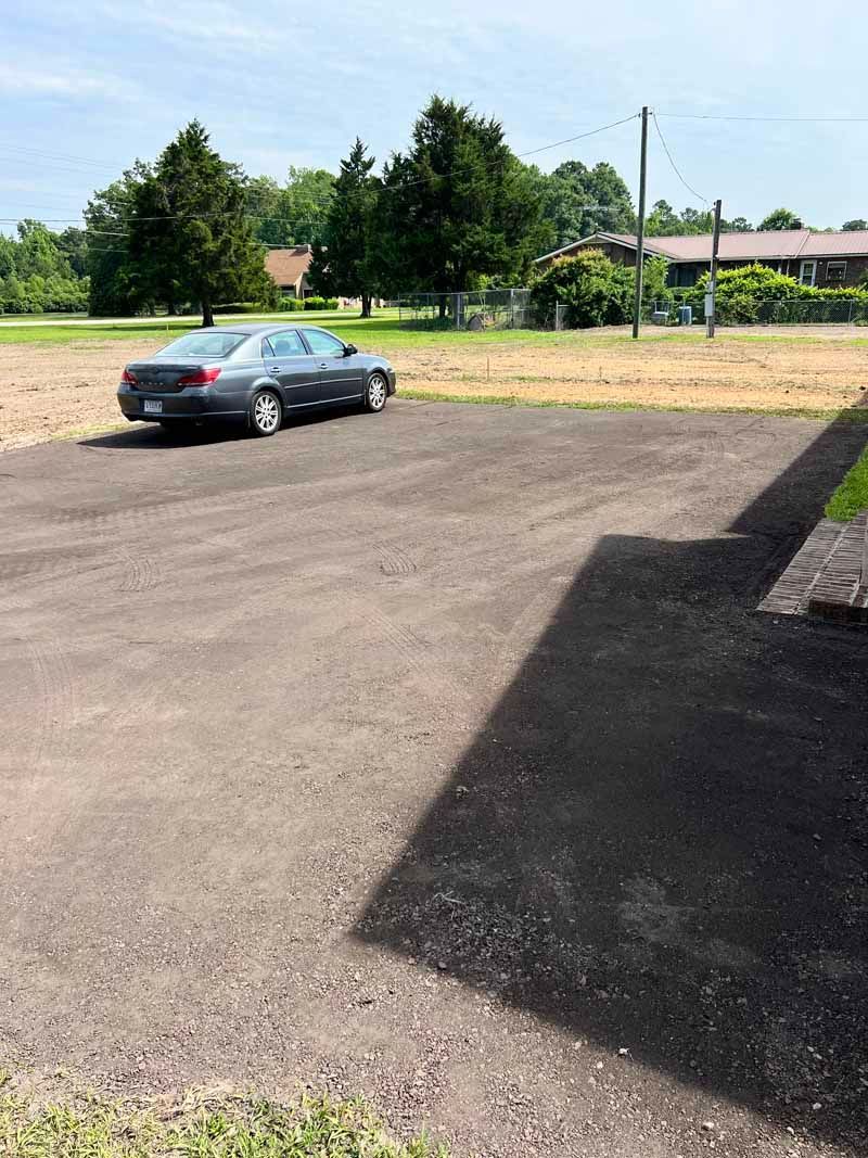 Silver sedan parked on a sunlit gravel lot beside trees and a building shadow