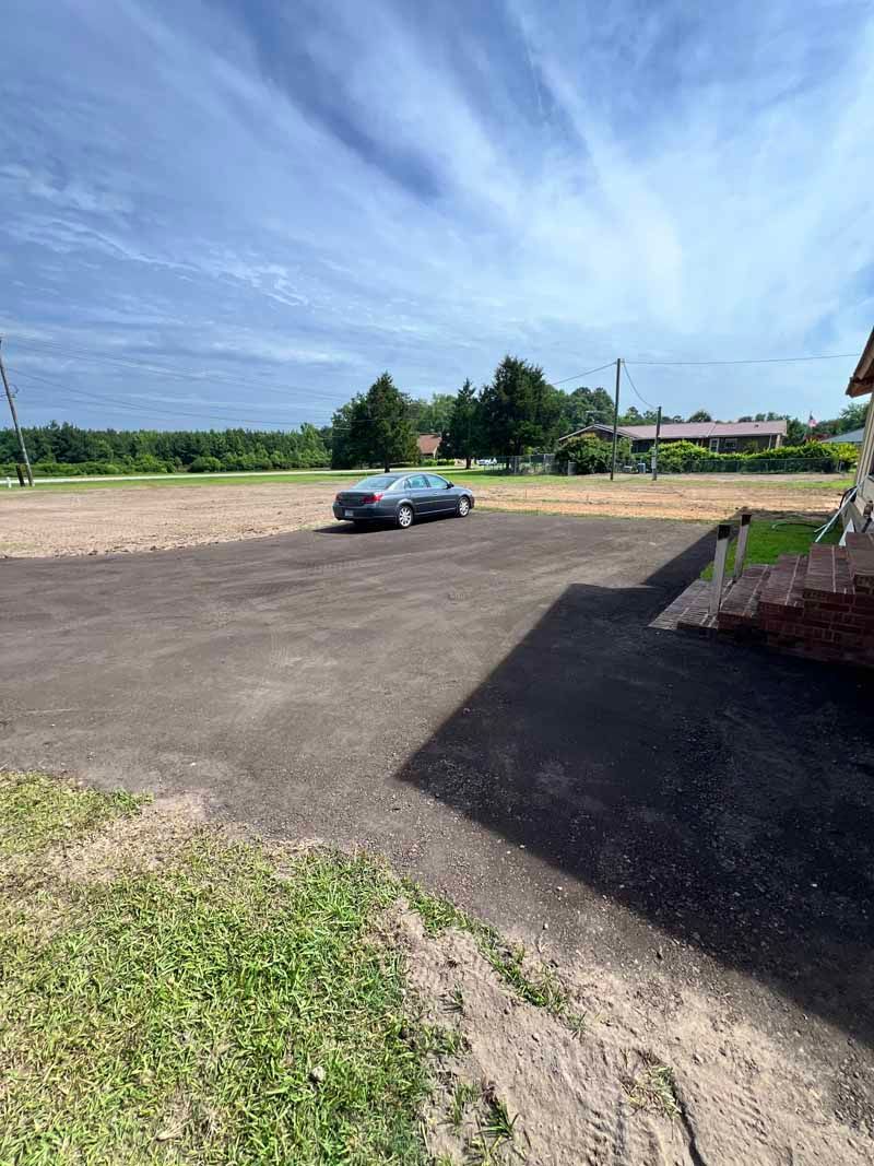 Blue car parked on a paved lot beside grassy fields under a bright blue sky