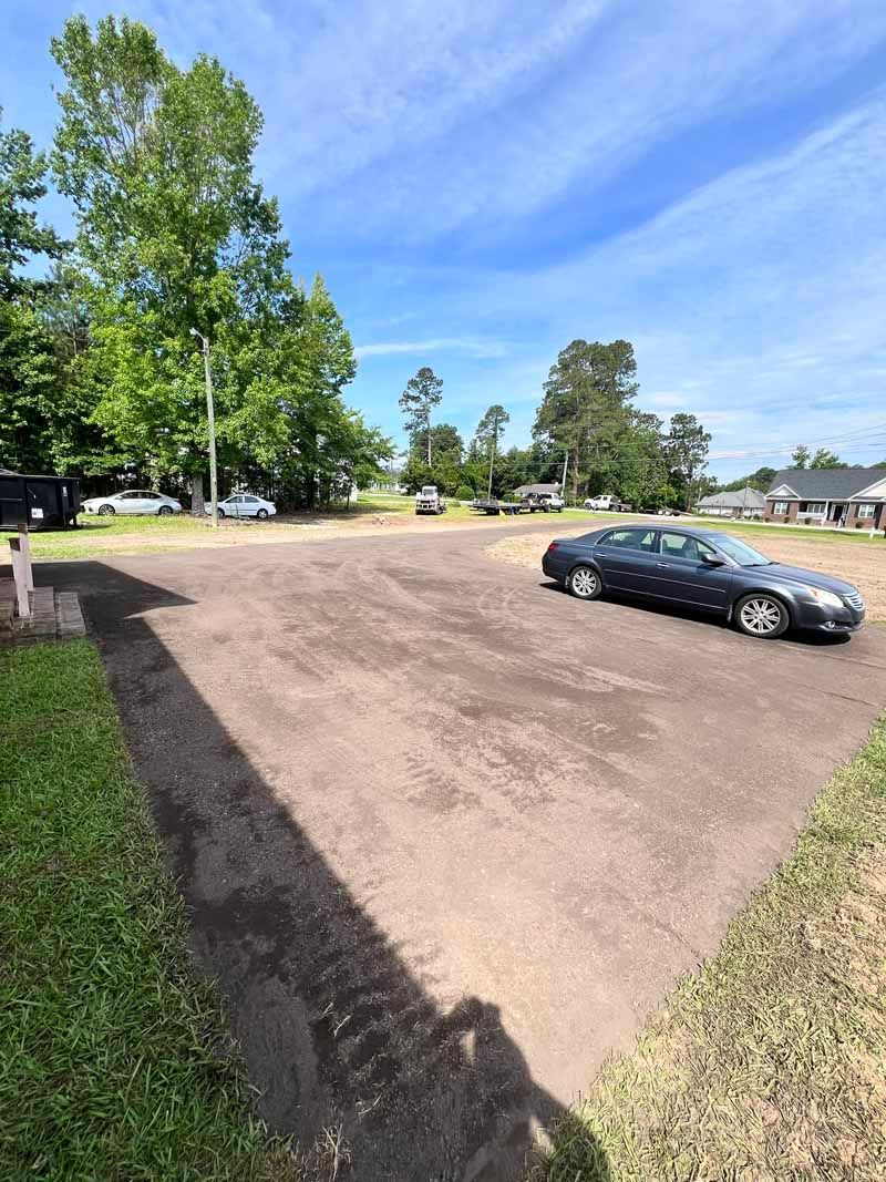 Gravel parking lot with a black car, tall trees, and a blue sky on a sunny day