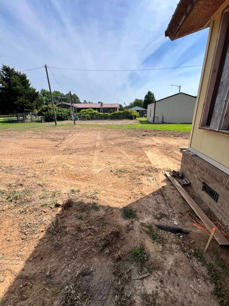 Empty dirt yard beside a house, with a shed and trees in the background.