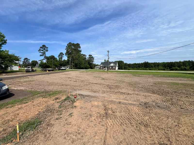 Empty dirt lot beside a road under a blue sky, with trees and power lines in the distance