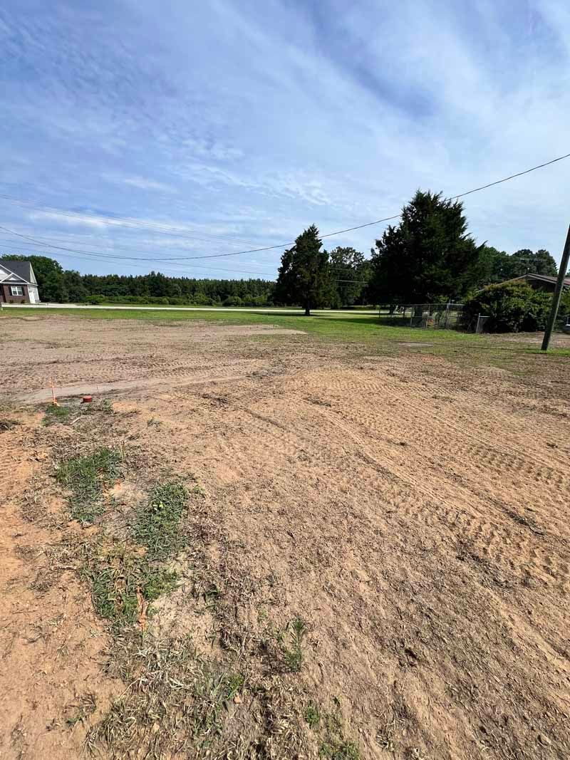 Bare dirt field with sparse grass, trees in the distance, and a cloudy blue sky