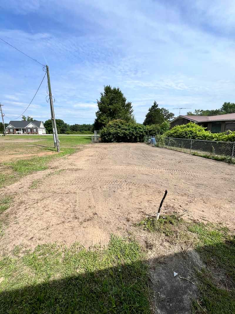 Bare dirt mound in a rural yard beside a grassy field, with a tree and houses under a blue sky.
