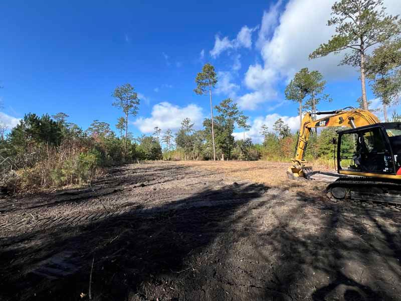 Cleared dirt road with a yellow excavator on the right under a blue sky and trees