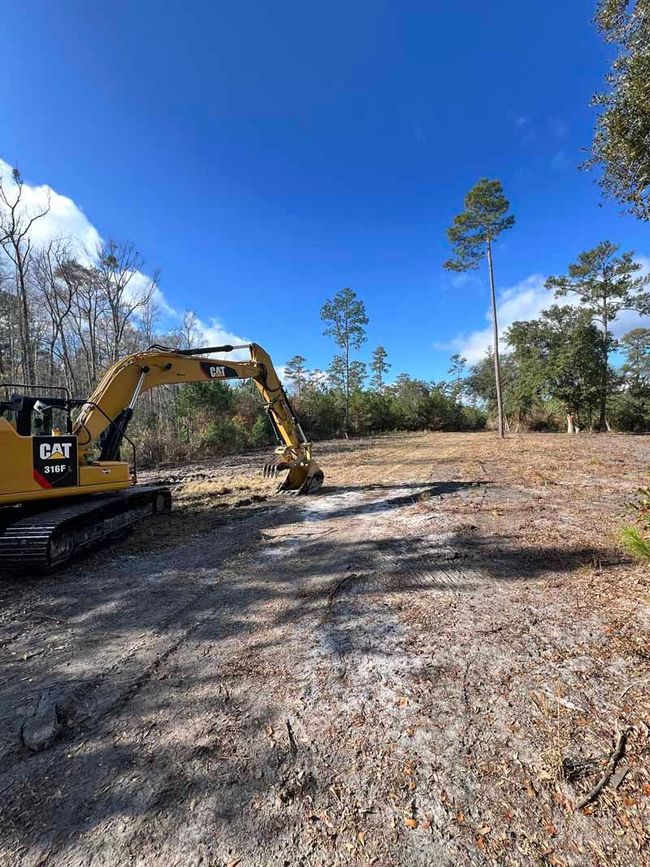 Yellow excavator clearing a dry, open lot under a bright blue sky with sparse trees
