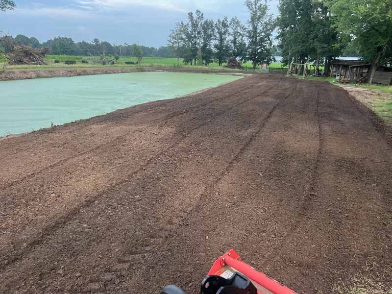 Freshly tilled soil beside a light blue pond, with a red tractor in the foreground and trees in the distance