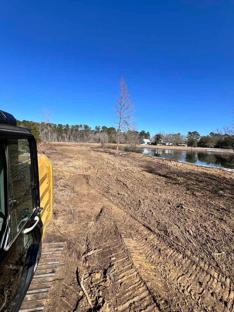 Bulldozer on muddy riverbank beside a calm pond under a clear blue sky