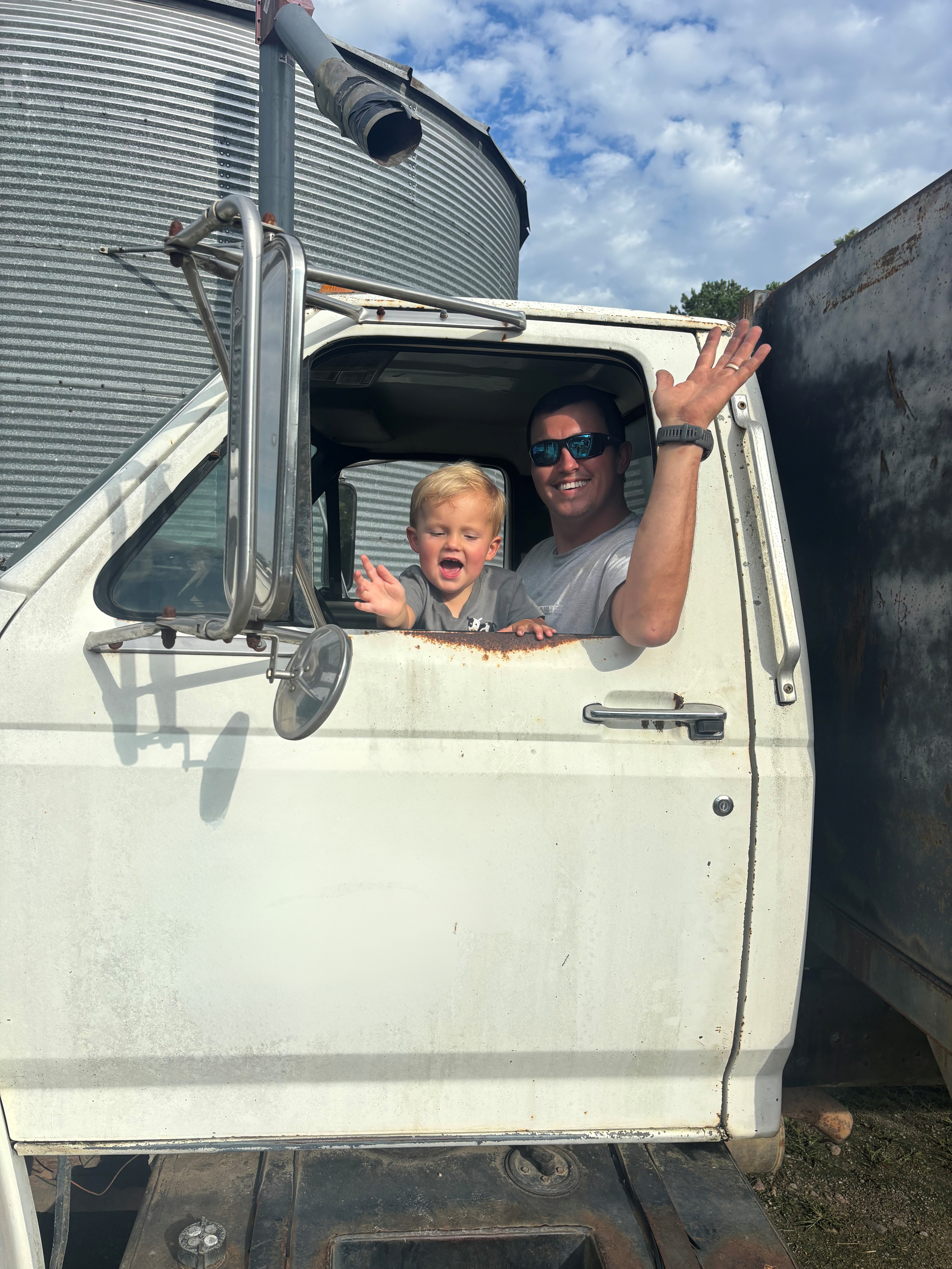 Two people wave from the cab window of a white truck parked beside a trailer in sunny weather