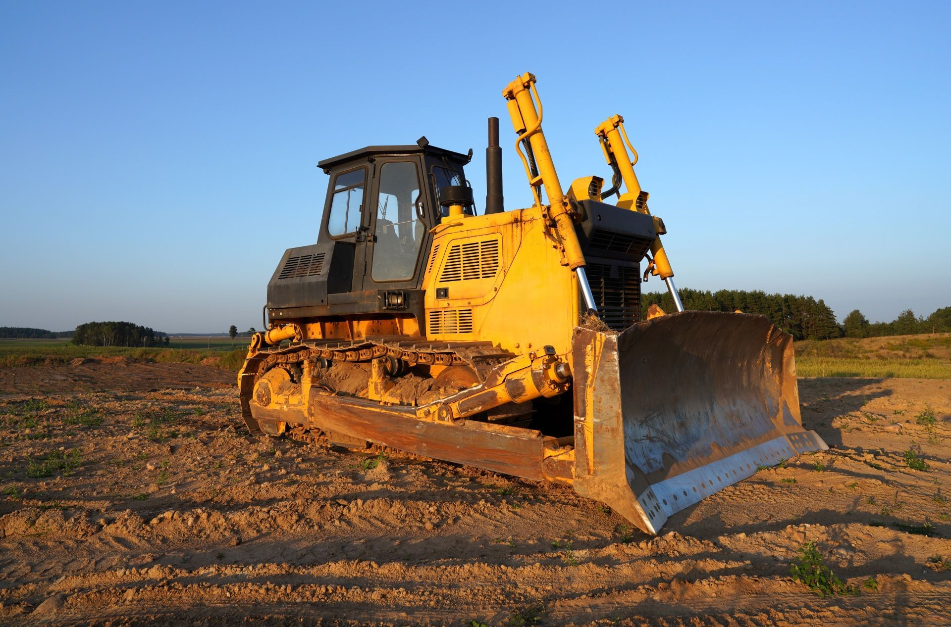 Yellow bulldozer on a dirt field at sunset, facing forward with raised blade.