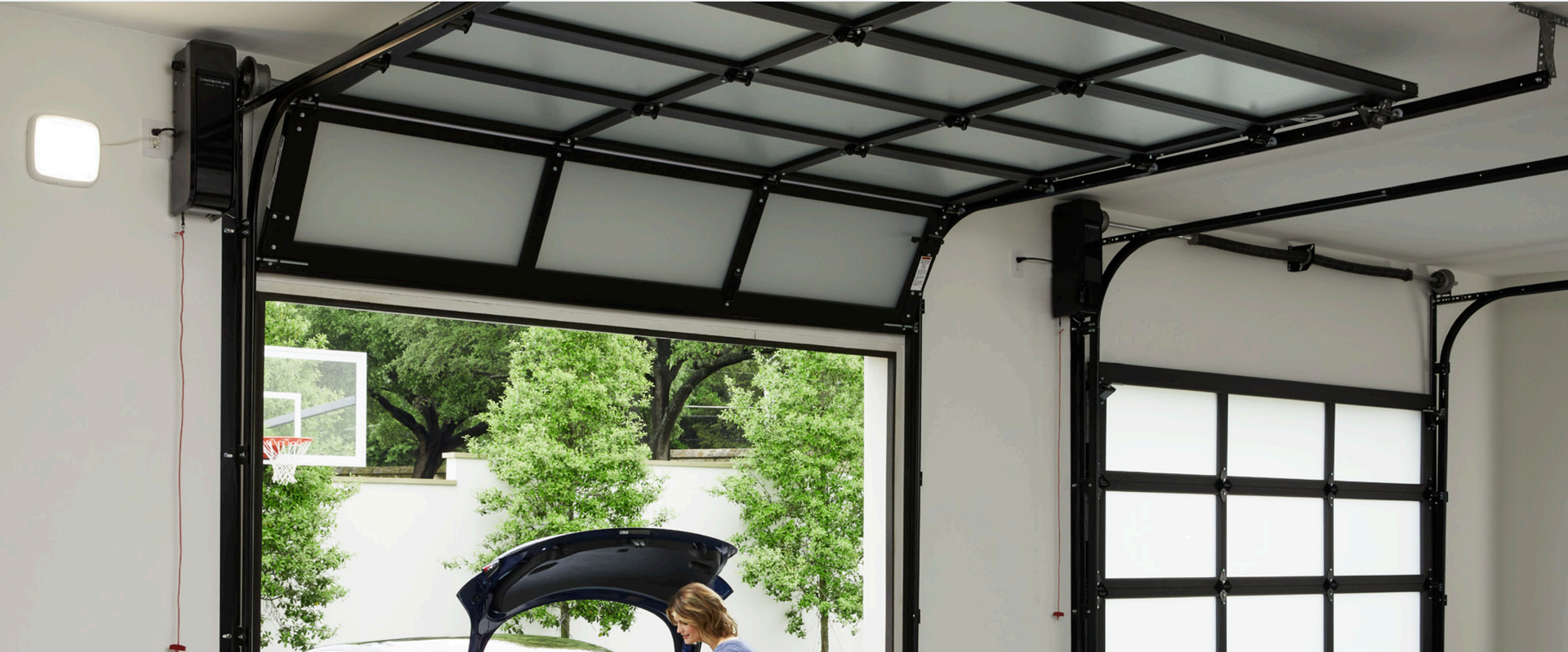 Garage door open, revealing a basketball hoop and trees. A person works on a car.