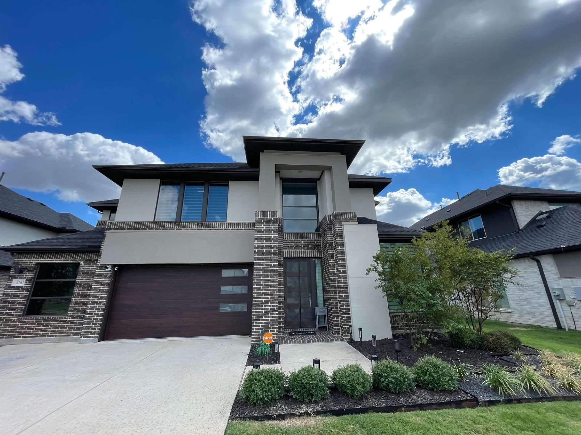Two-story modern home with gray stone and stucco exterior, brown garage door, and blue sky.