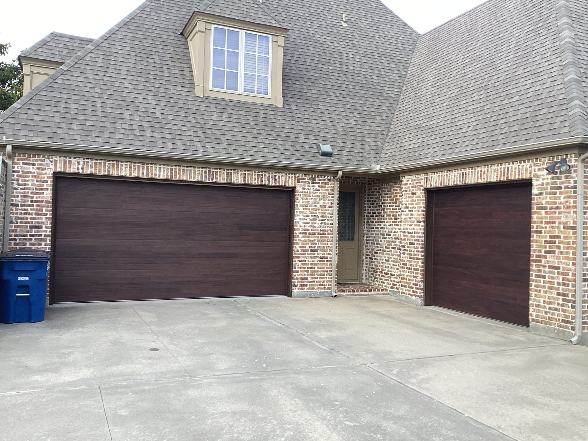 A brick house with two garage doors and a blue trash can