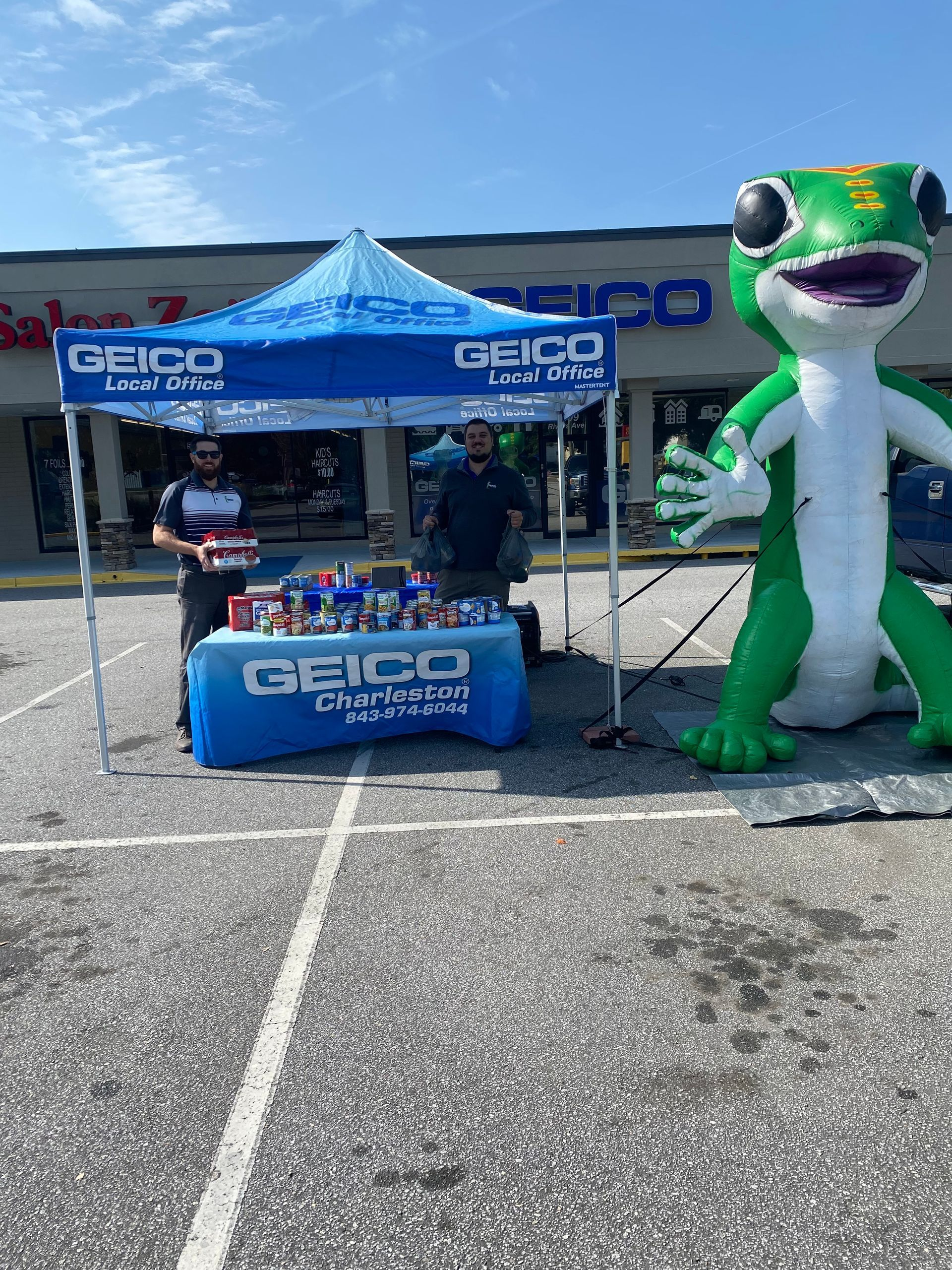 Geico booth with two men and a large gecko inflatable outside a building with the Geico logo. Blue tent.
