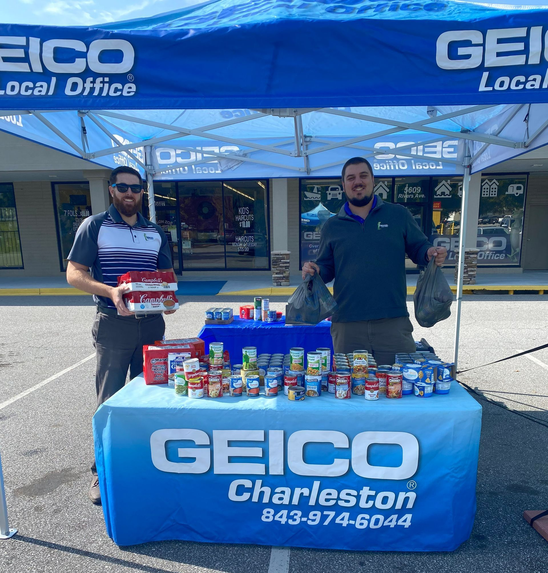 Two men stand behind a GEICO table with canned food, outside a Charleston business.