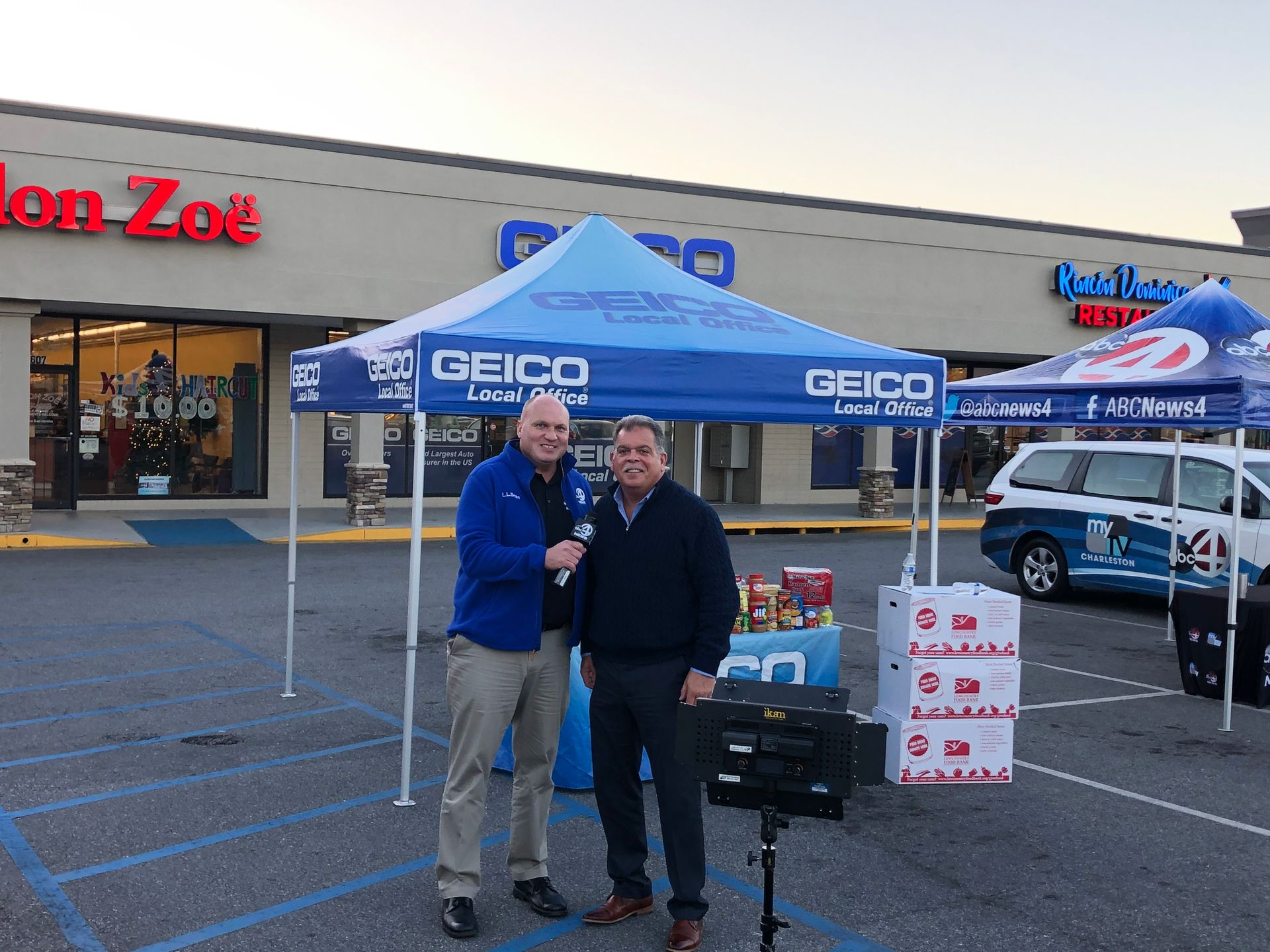 Two men at a Geico promotional event outside a store. One holds a microphone, the other smiles.