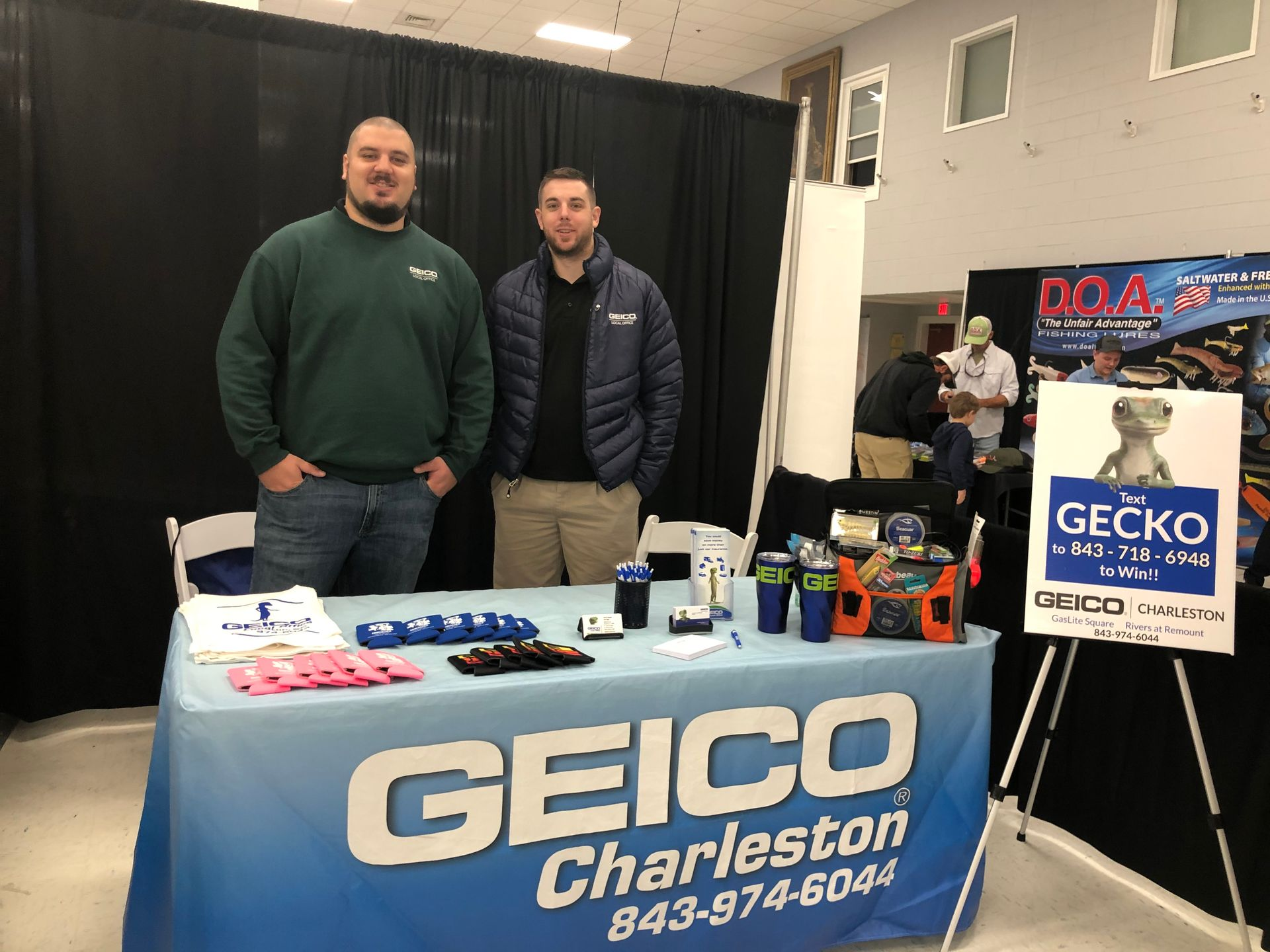 Two men at a GEICO Charleston booth. One wears a green shirt, the other a blue jacket. Booth has promotional items.