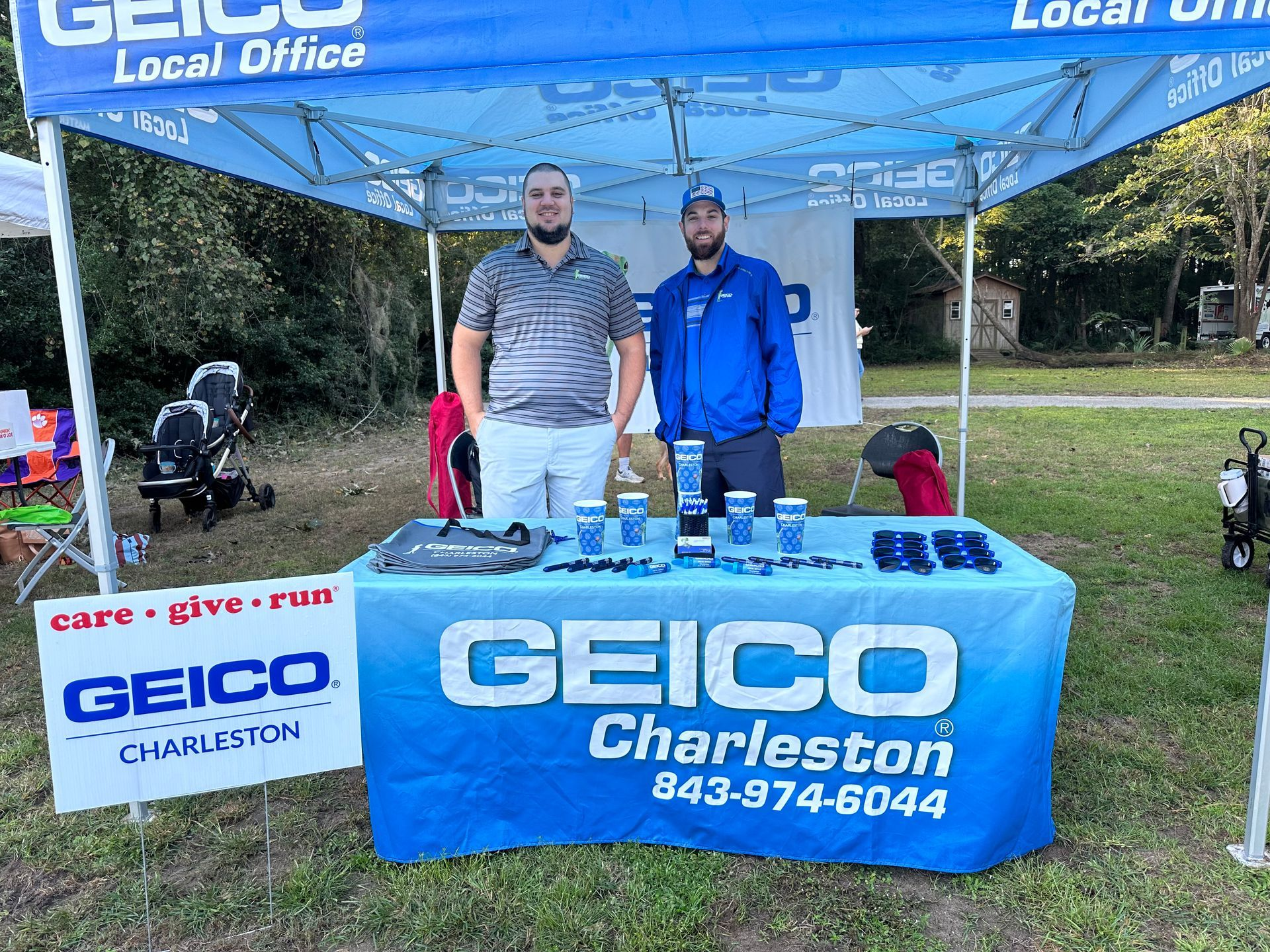 Two men at a GEICO booth in Charleston. The booth has the company logo, and they are surrounded by promotional items.