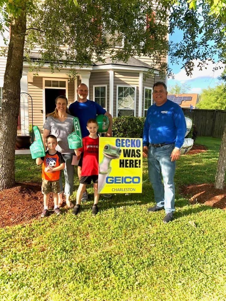 Family poses with a GEICO sign in front of a house. A man stands at the right.