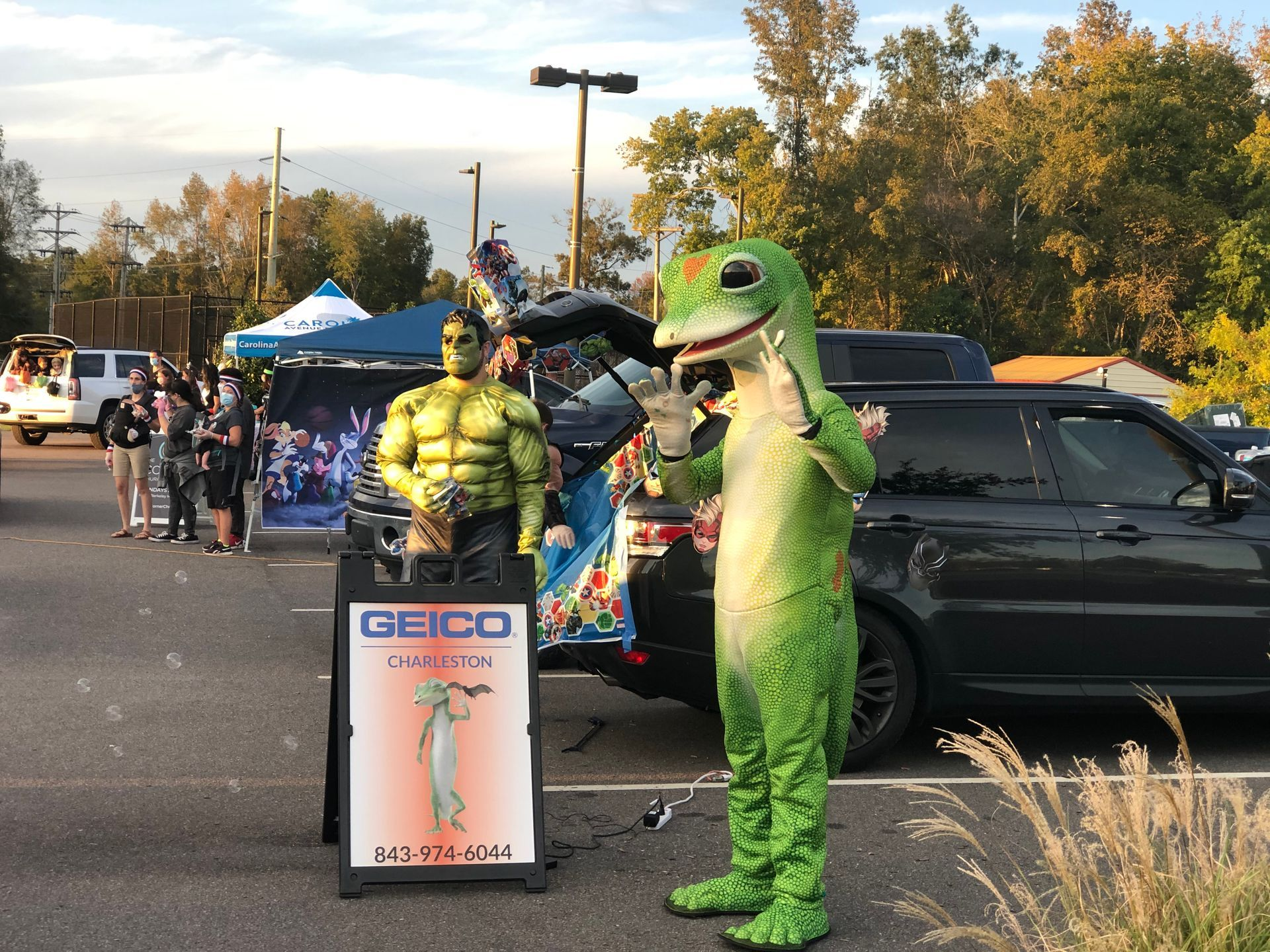 Geico gecko mascot at a promotional event, holding a microphone near a sign with the Geico logo.
