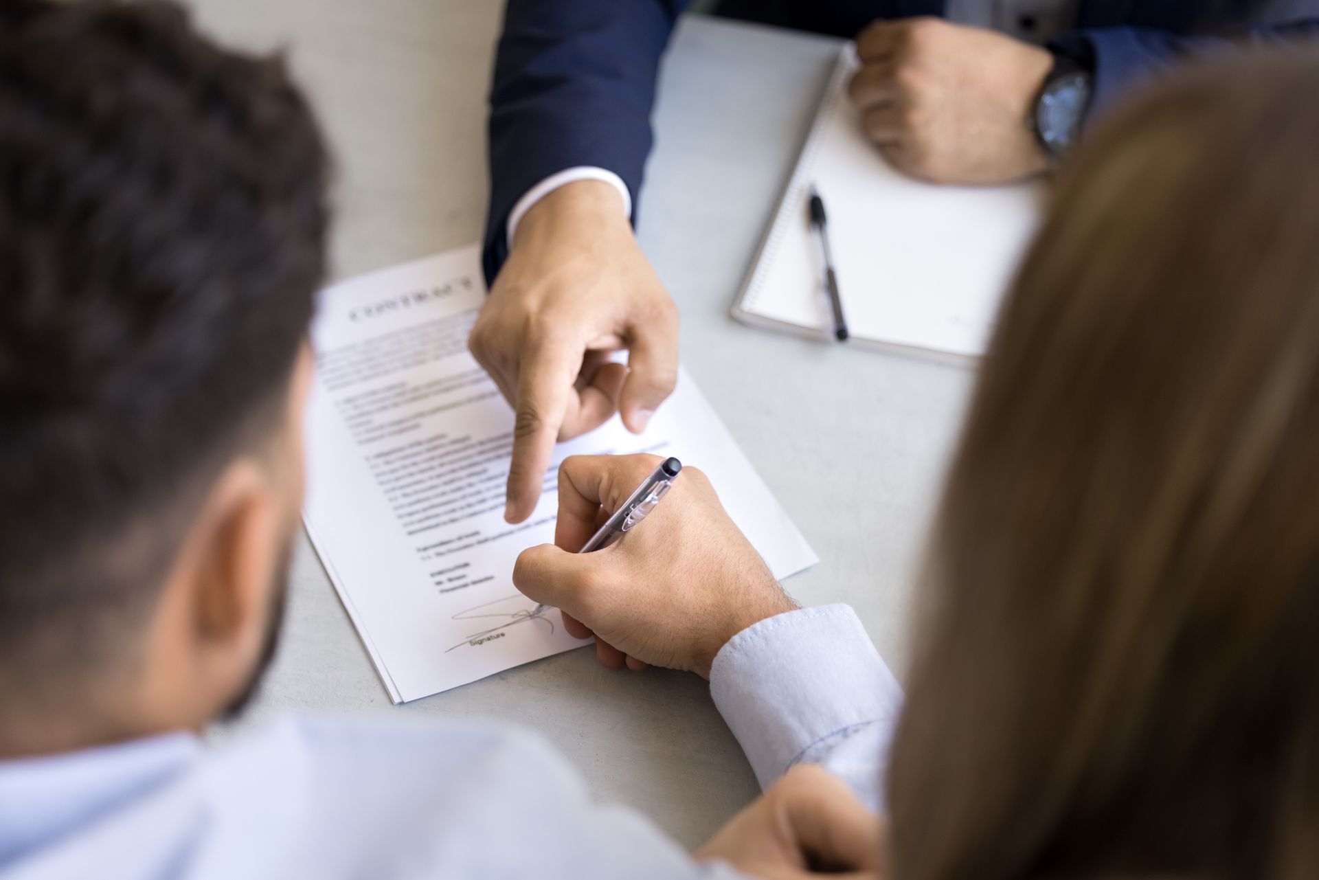 Person signing a contract, guided by another person's pointing finger, at a table.