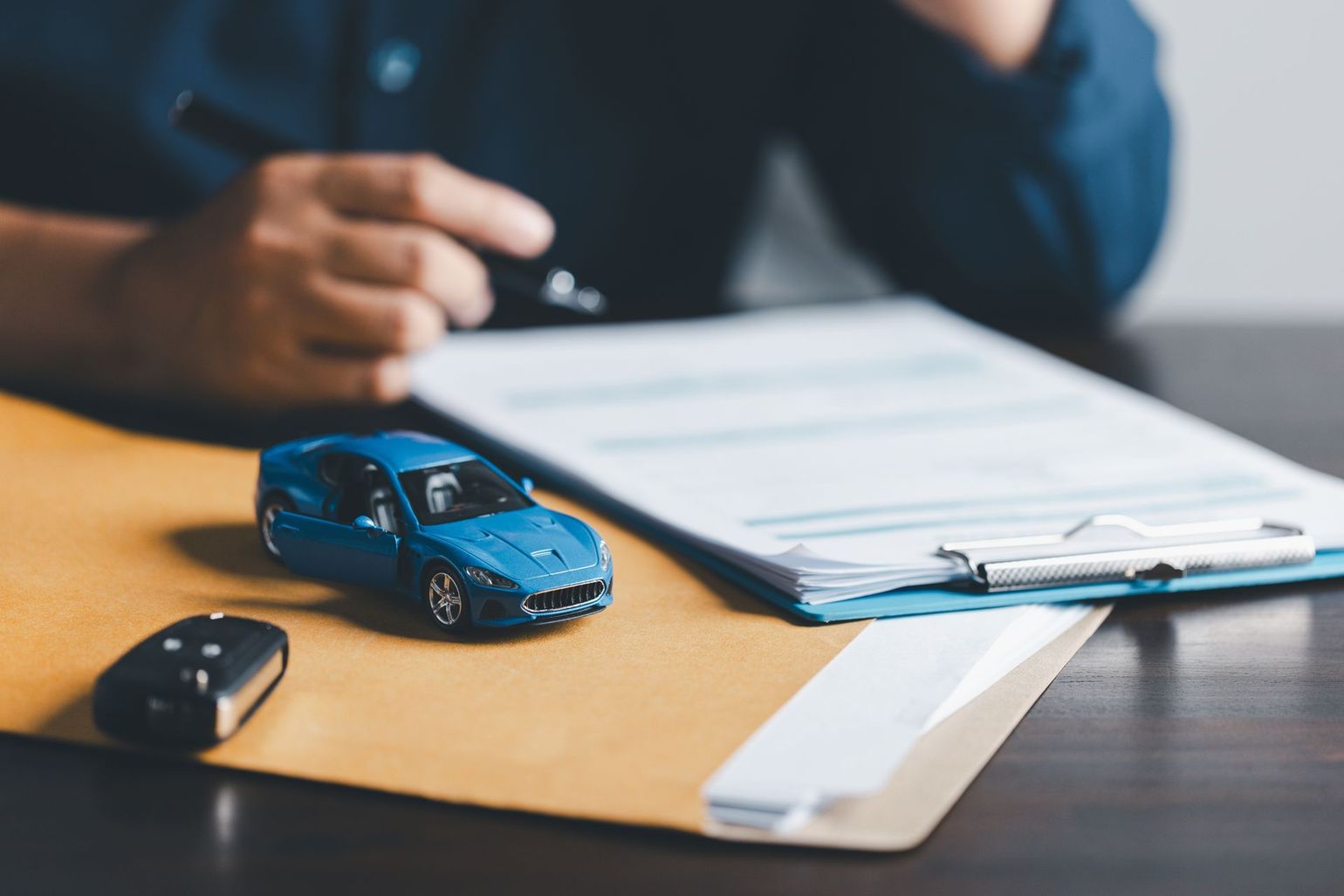 Person with a pen reviewing car insurance paperwork next to a toy car and car keys on a desk.