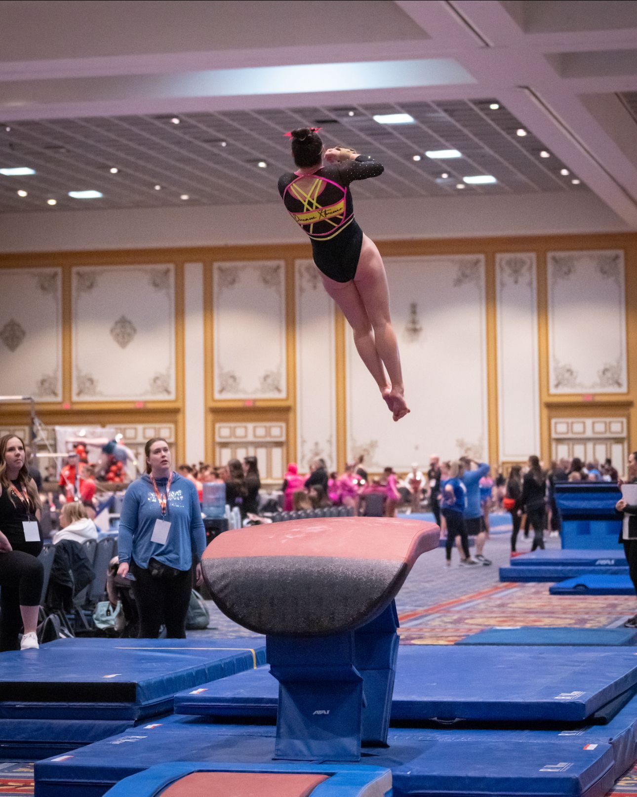 A female gymnast is jumping over a balance beam in a gym.