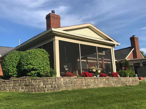 Screened porch with brick chimney, surrounded by green grass and a stone retaining wall. Red flowers and shrubs.