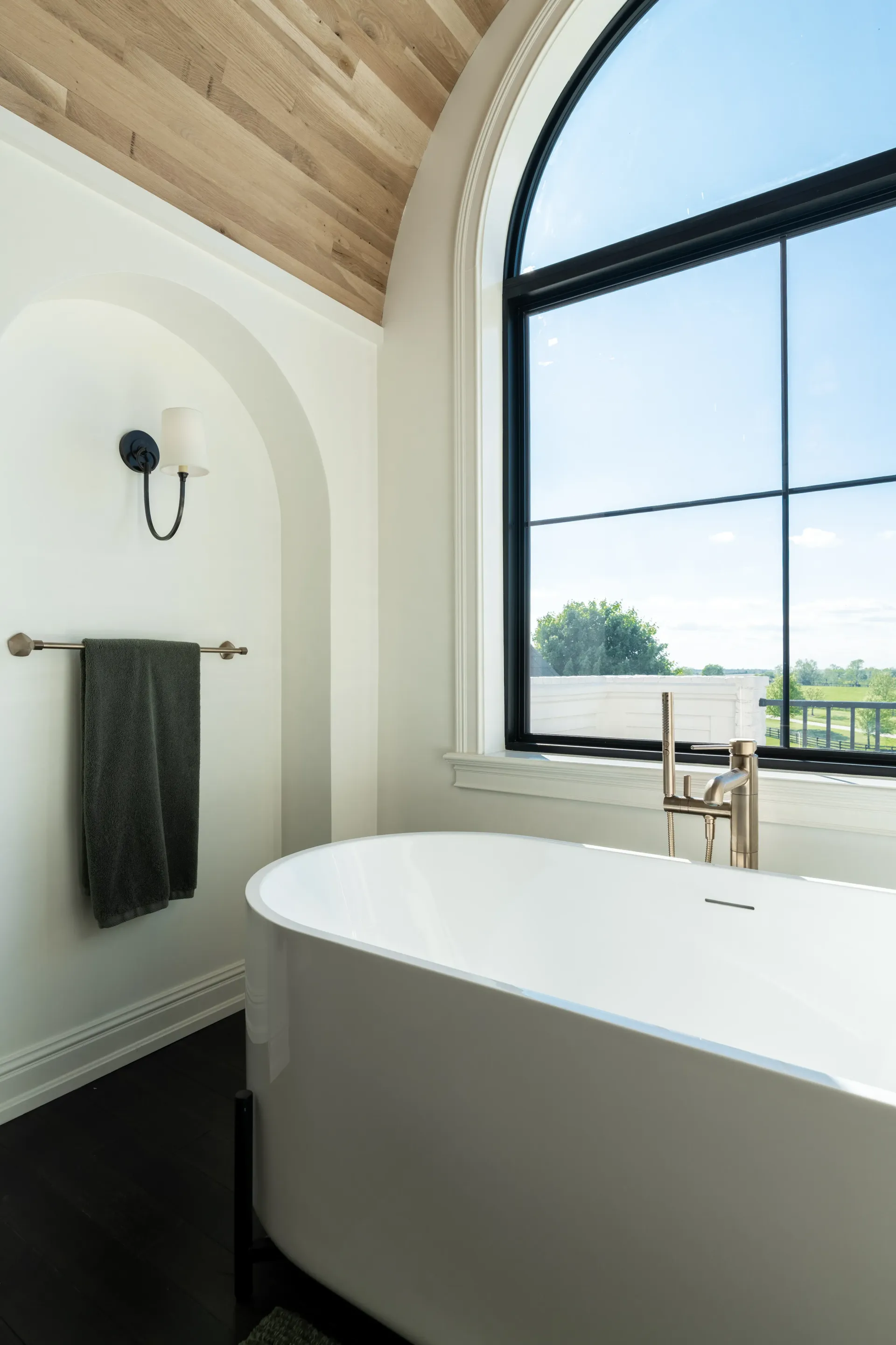 Bathroom with freestanding white tub, large arched window, towel on a rack, and a wood-plank ceiling.