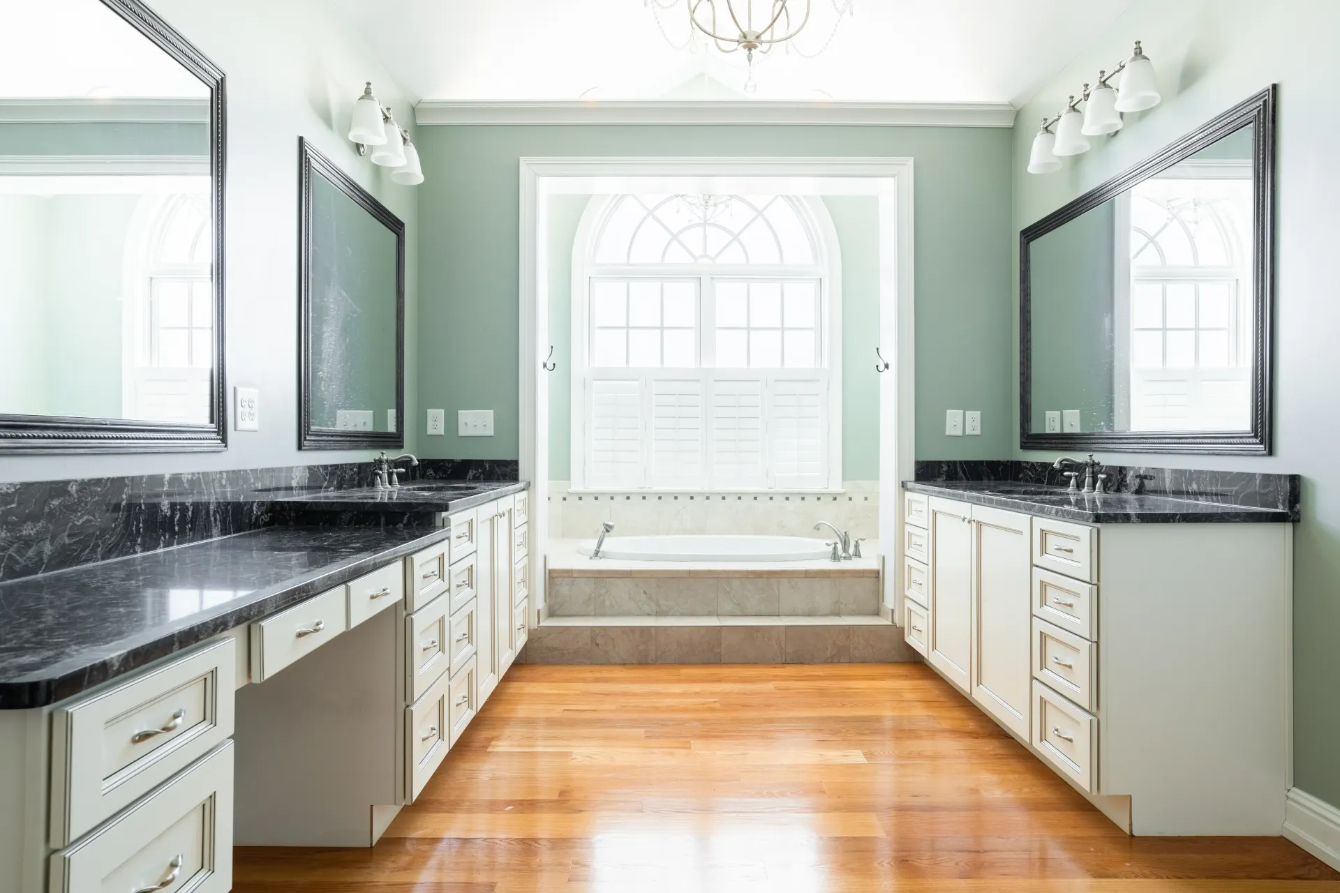 Spacious bathroom with dual vanities, large mirrors, and a sunken bathtub, hardwood floor, light blue walls.