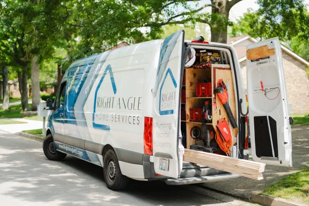 White work van with open doors, displaying tools and lumber. Parked on a street with trees and a house.