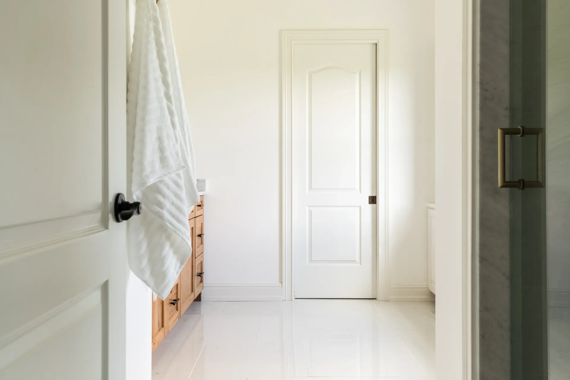 Bathroom interior with white walls, door, and floor; towel hanging on a door, wooden vanity.