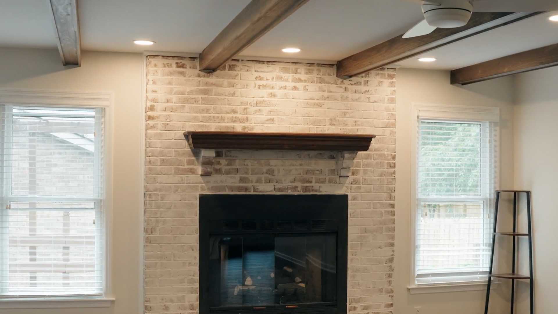 Fireplace with brick surround, dark mantel, and wooden beams. Two windows flank the fireplace.