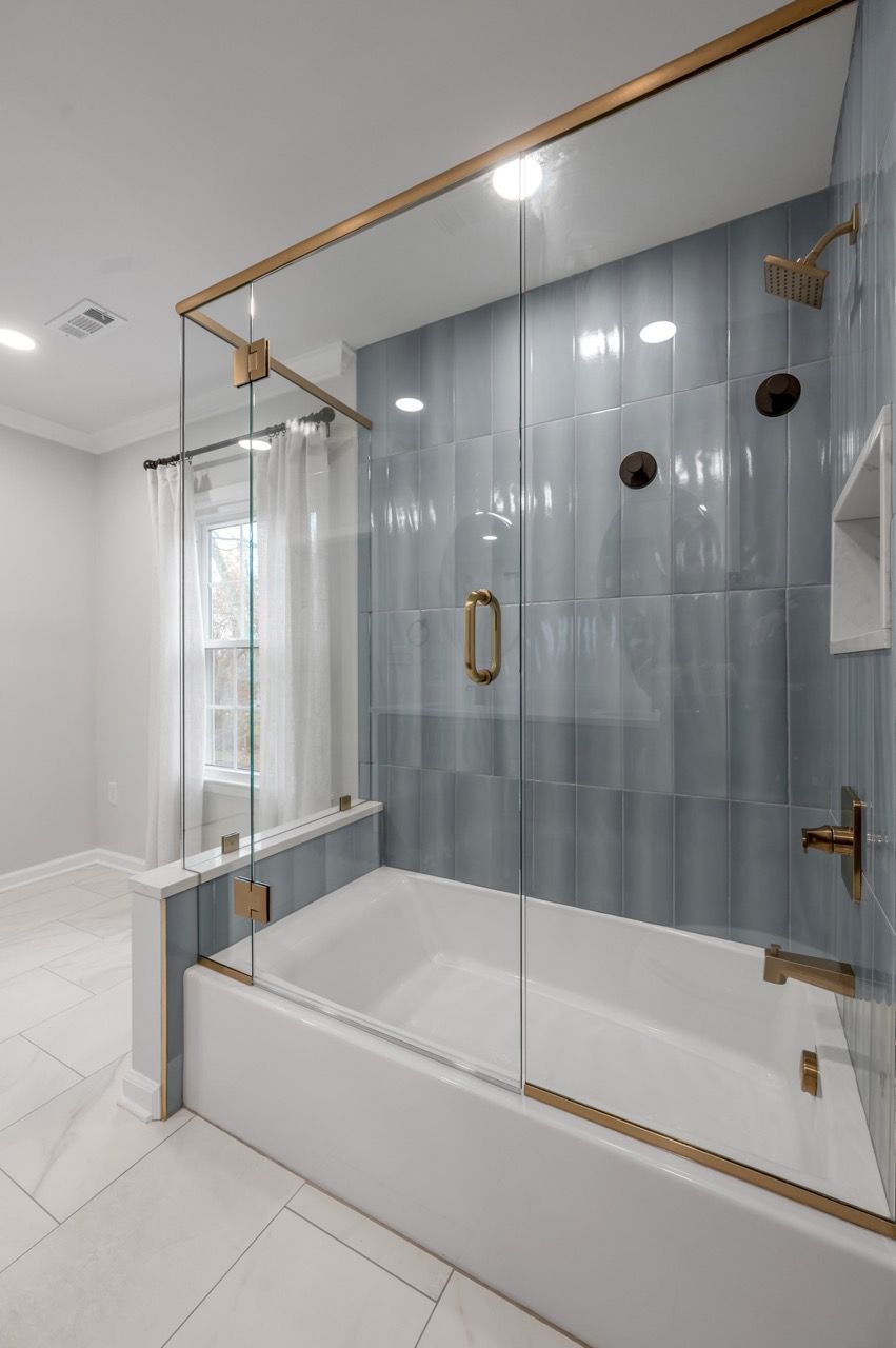 Bathroom with blue tiled wall, white tub, and glass shower enclosure with gold accents.