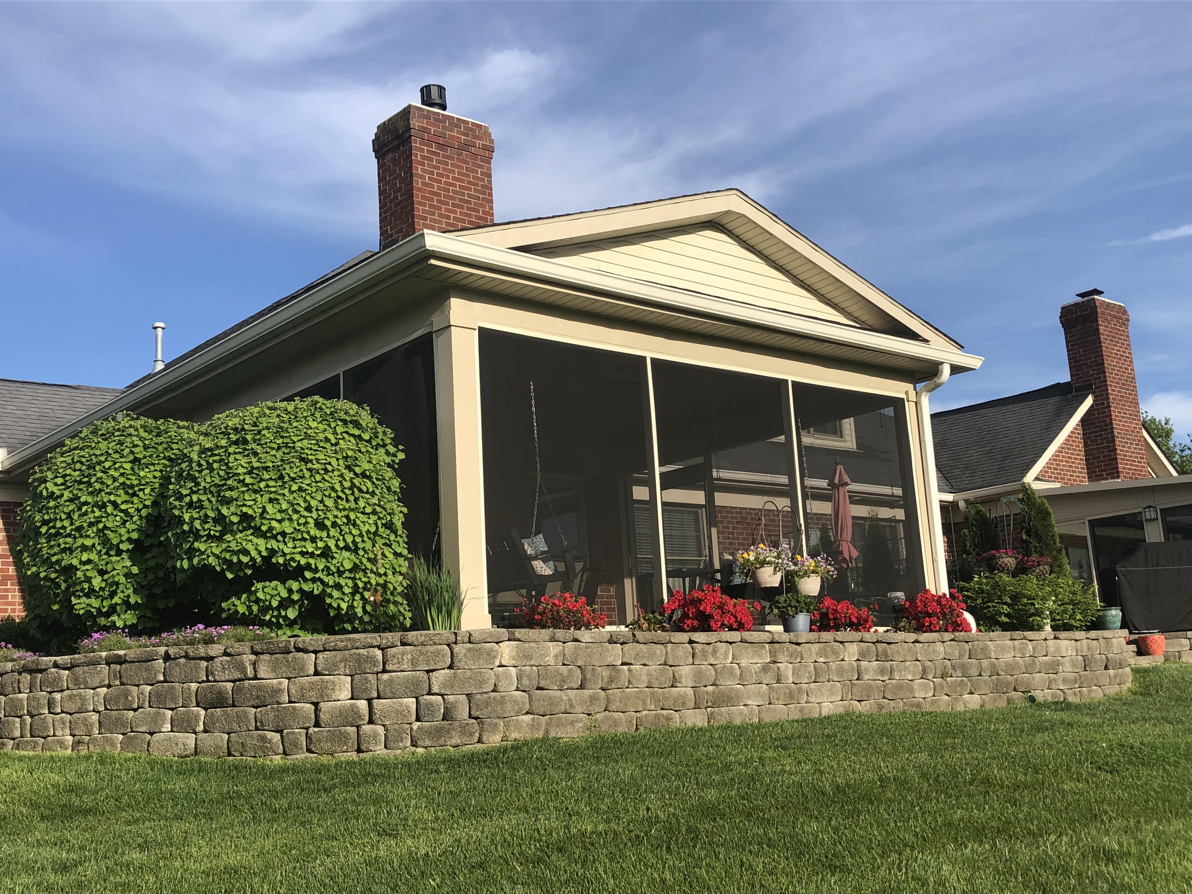 Screened-in porch with brick chimney. Red flowers, green shrubs, and a low stone wall are visible.