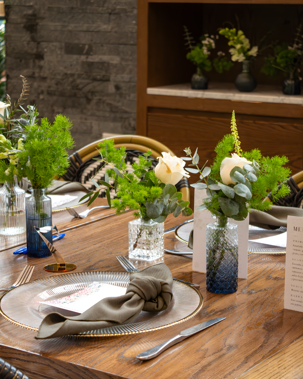 Mesa de comedor rústica con centros de mesa florales blancos, cristalería y platos puestos para una comida.